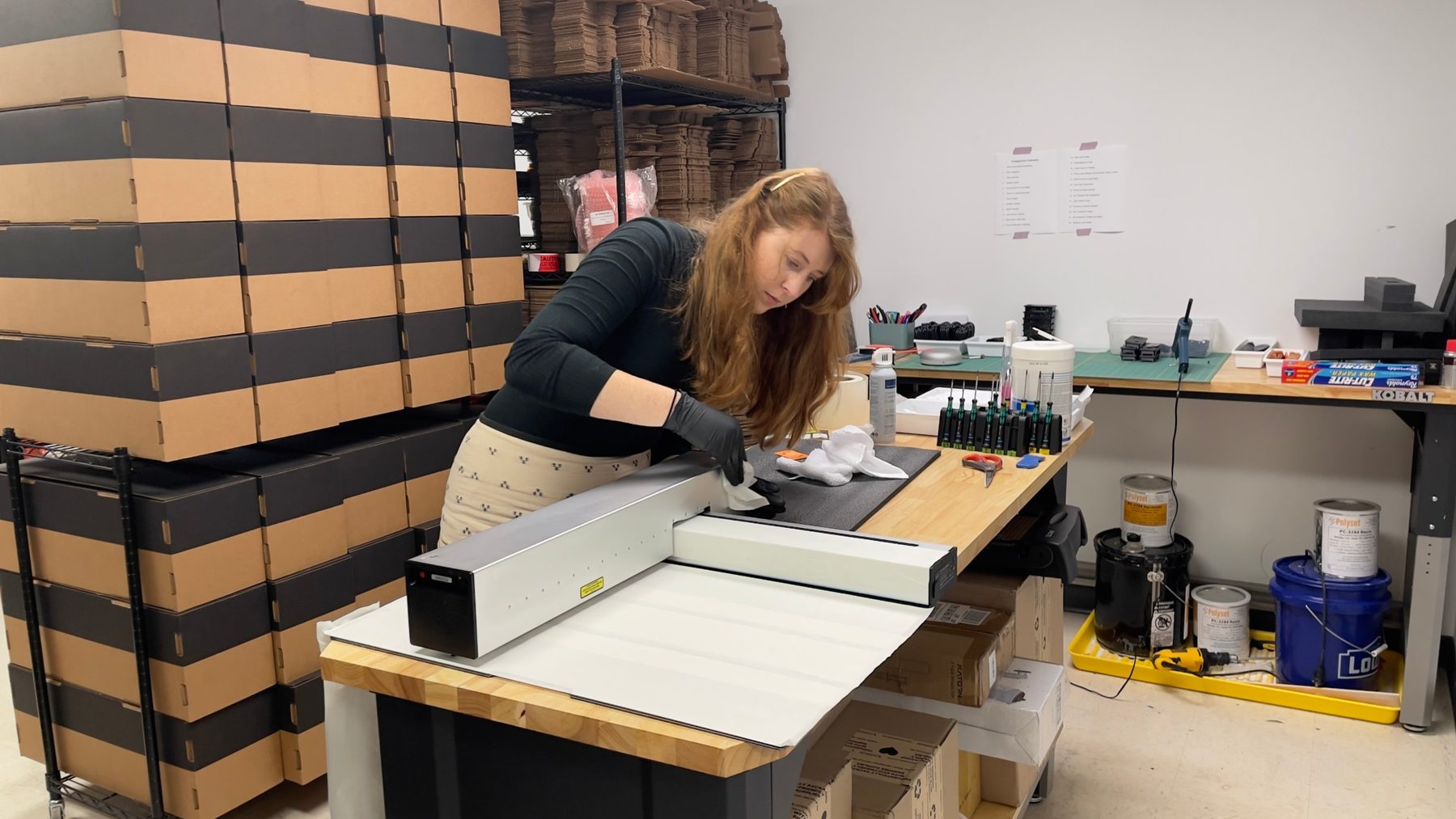 Sophie cleaning a tested Optic at the packing bench, stacked shipping boxes behind her